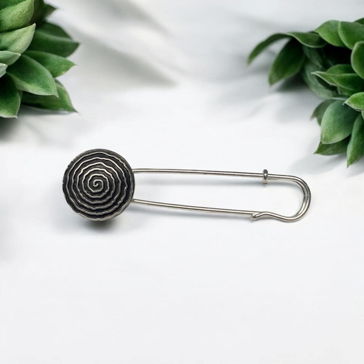 Ponchonadel mit rundem Spiralmuster in Altsilber-Optik auf weißem Untergrund vor grünen Blättern