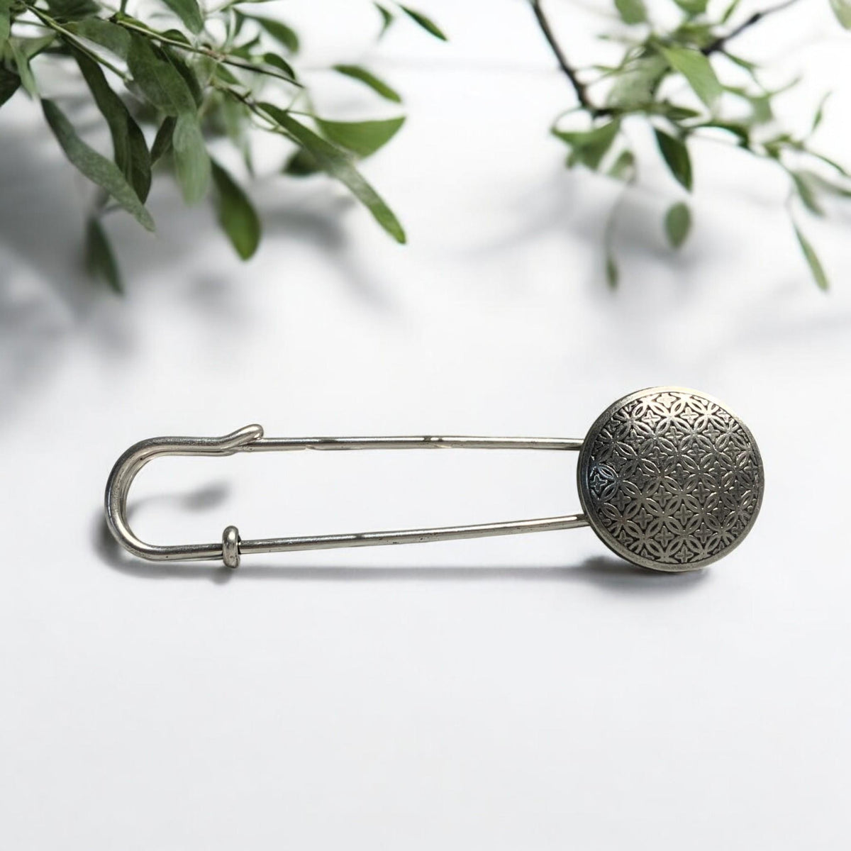 Ponchonadel mit Blume des Lebens in Altsilber-Optik auf weißem Untergrund vor Grünpflanzen