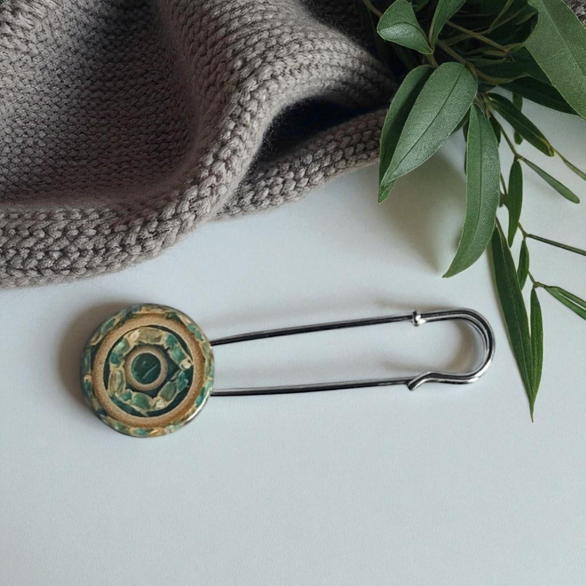 Ponchonadel mit rundem Holzelement in Beige-Grün neben grauem Strick und Zweigen