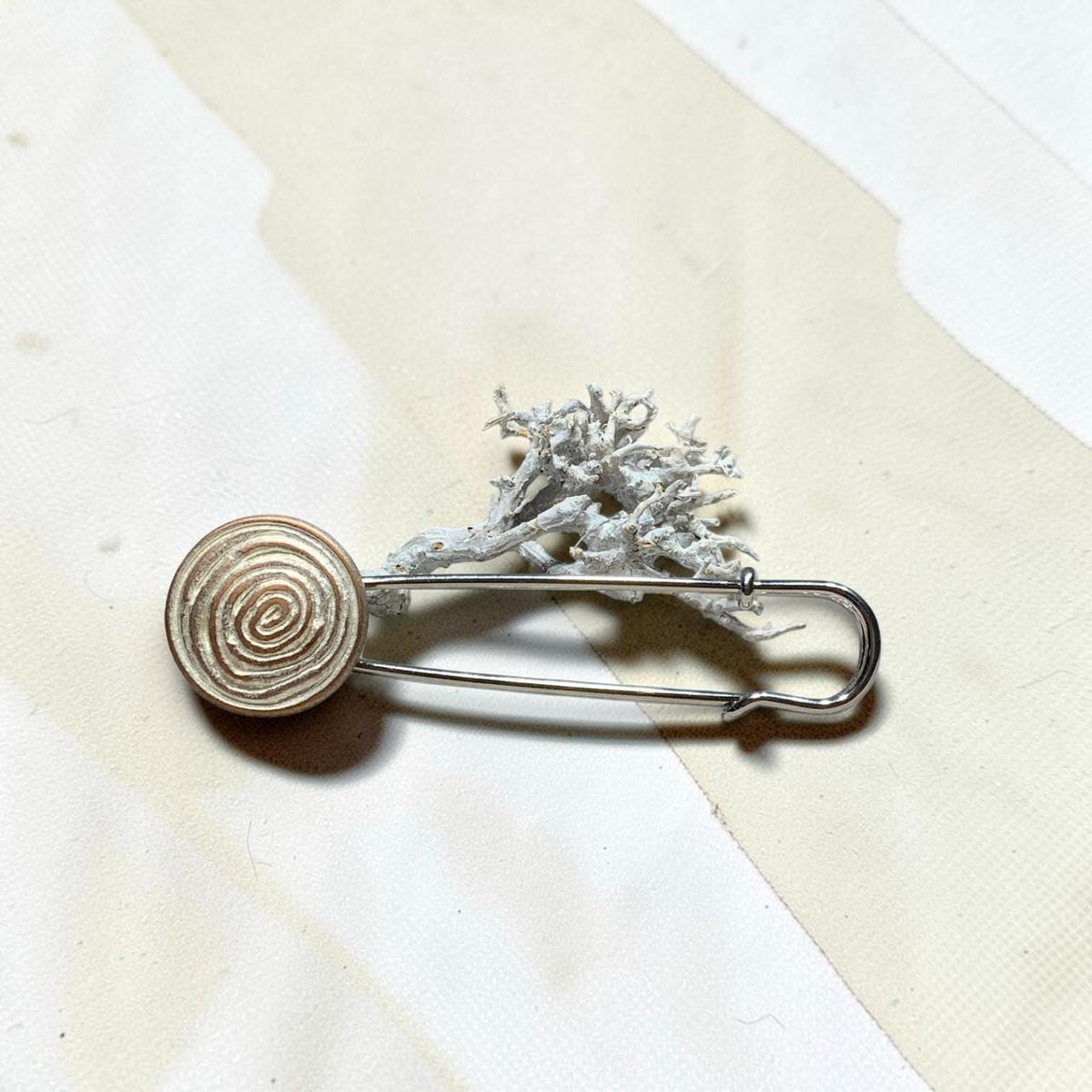 Ponchonadel mit hellem Spiralmotiv auf cremeweißem Untergrund vor Trockenflechte